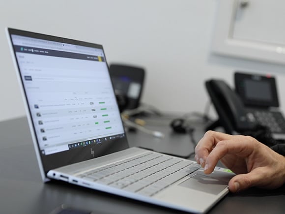 A person using a silver laptop at a desk, viewing a webpage displaying sales or inventory data, with office phones in the background.