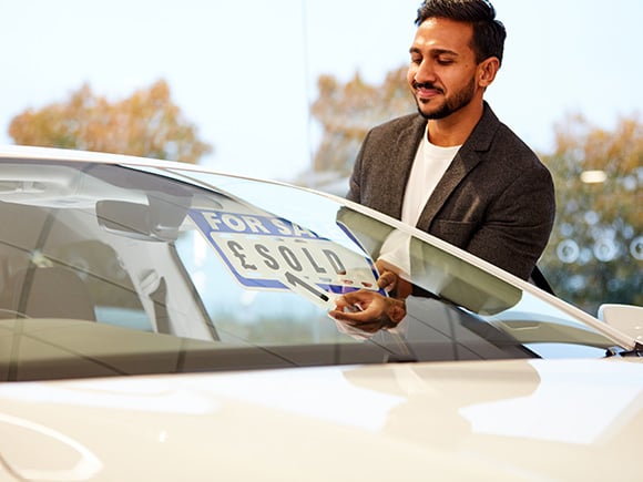 A person in a blazer places a "SOLD" sign on the windscreen of a white car inside a showroom, indicating the vehicle has been purchased.