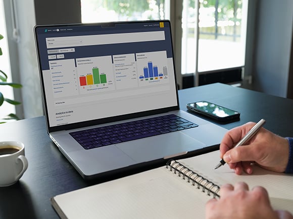 A person writes in a notebook while looking at performance graphs on a laptop screen, with Auto Trader Intelligence software open, on a desk next to a phone and a cup of coffee.