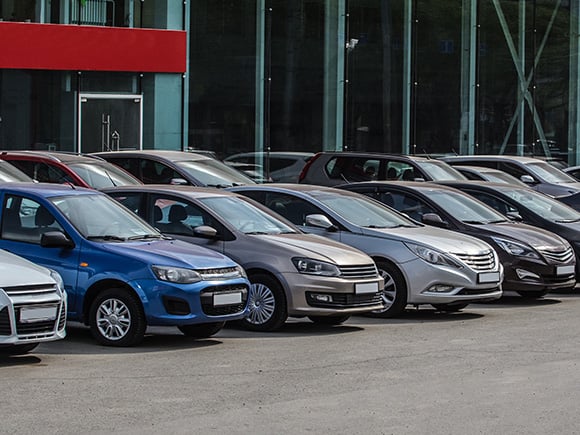 Row of parked cars in front of a glass-fronted dealership building, featuring a variety of makes and colours.