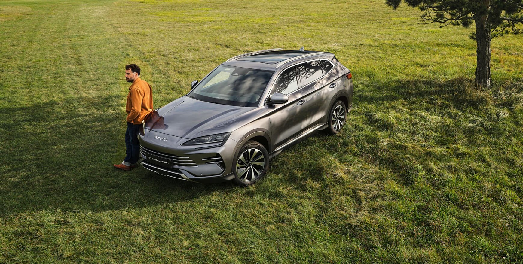 A BYD Seal U DM-i SUV parked on a grassy field with a man in a mustard-colored jacket standing beside it, looking into the distance.